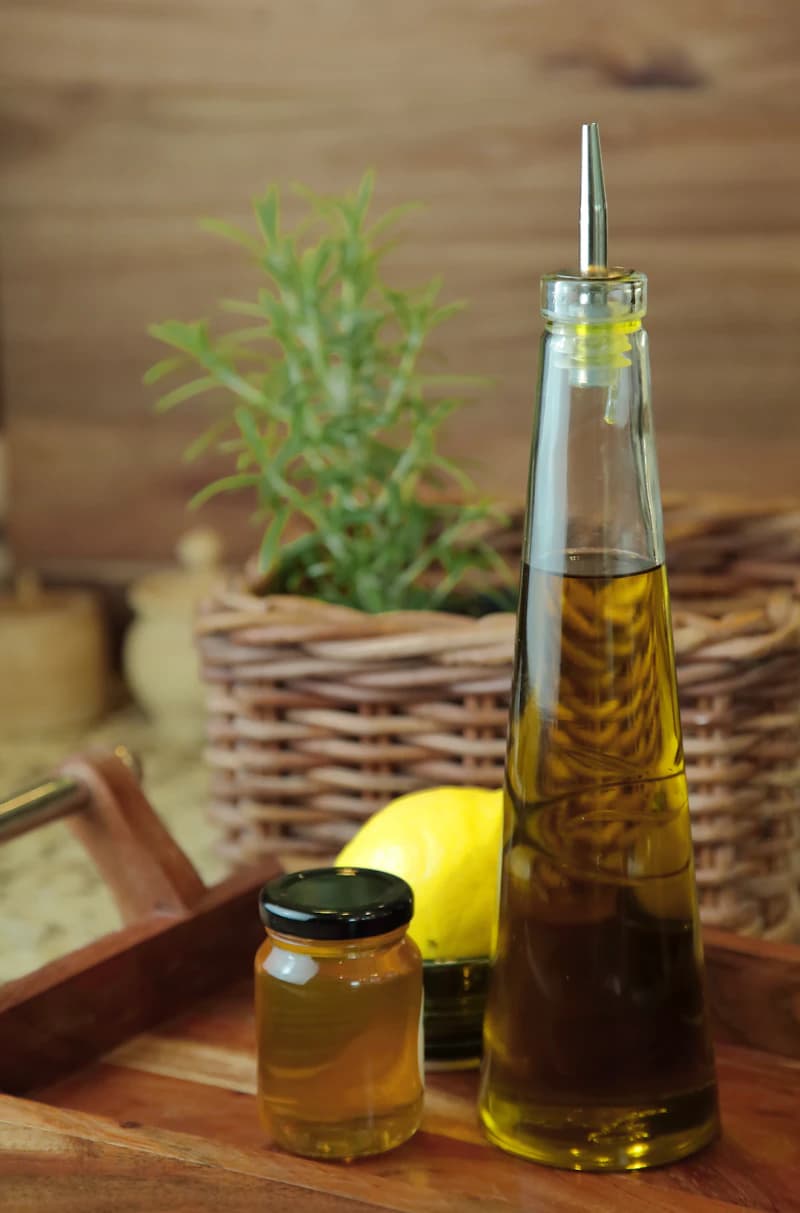 Une bouteille d’huile d’olive près de miel, de citron et de romarin sur un plateau en bois.